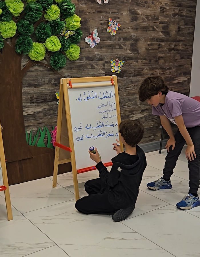 Two Arabeez students practising Arabic writing at easels in front of the classroom tree